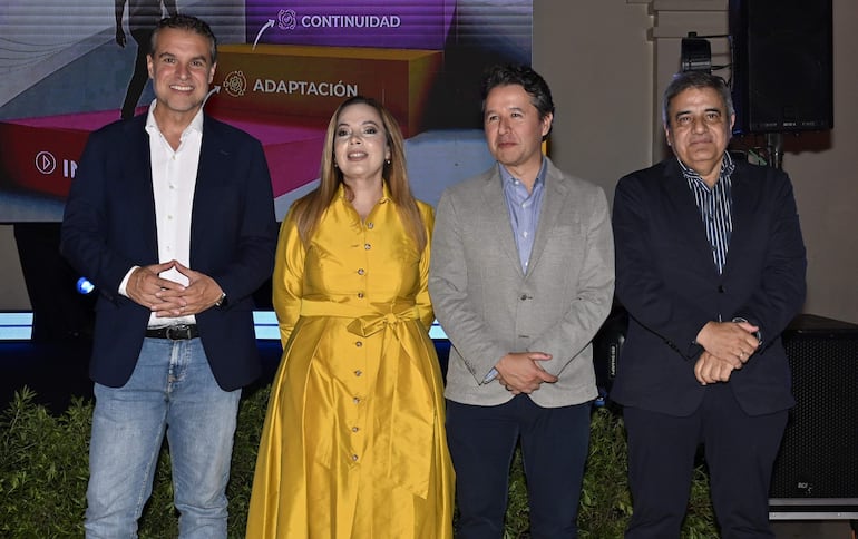 Cuatro personas sonríen en evento formal: hombre con chaqueta oscura, mujer en vestido amarillo, hombre en abrigo claro y otro en abrigo oscuro.