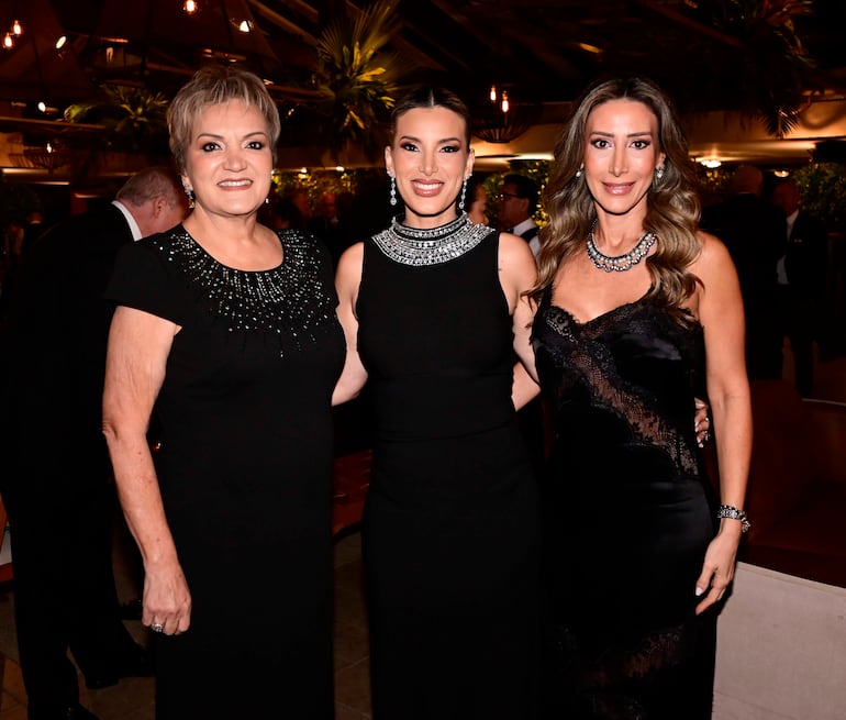 Tres mujeres en vestidos negros sonríen. Un ambiente iluminado con plantas decorativas alrededor.