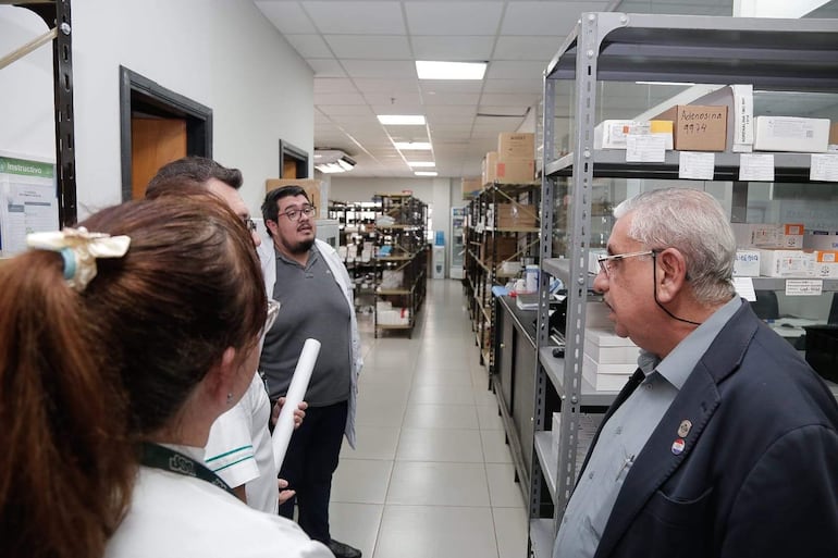 Isaías Ricardo Fretes, vestido formalmente, conversa con un hombre joven en laboratorio, rodeado de personas escuchando atentamente.