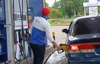 Hombre con camiseta azul y blanca y gorro rojo, llena su auto azul en estación de servicio con vegetación al fondo.