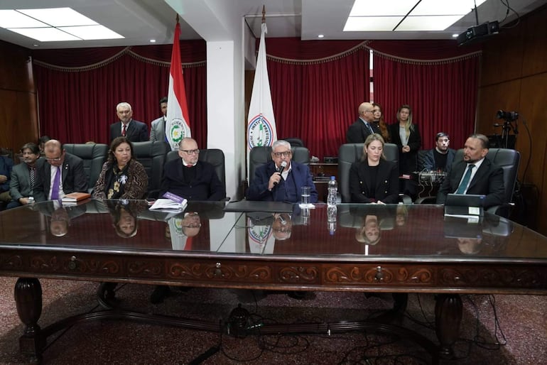 Isaías Fretes en traje gris explica, acompañado de mujeres y hombres en un ambiente formal, con banderas al fondo.
