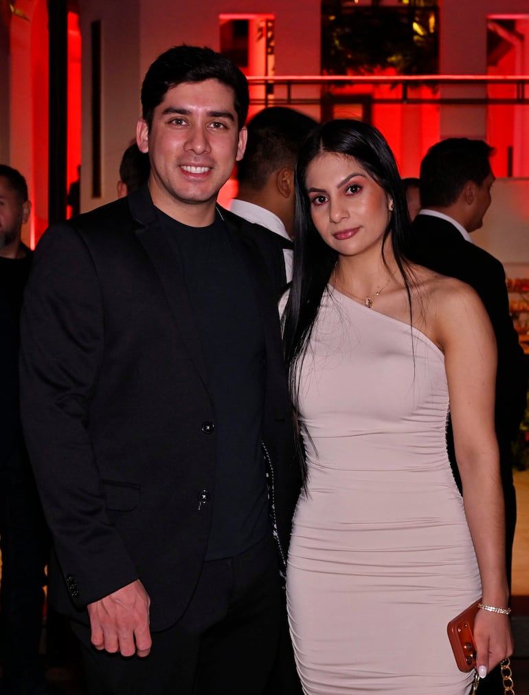 Hombre en traje oscuro sonríe junto a mujer en vestido nude, en un evento social con iluminación cálida.