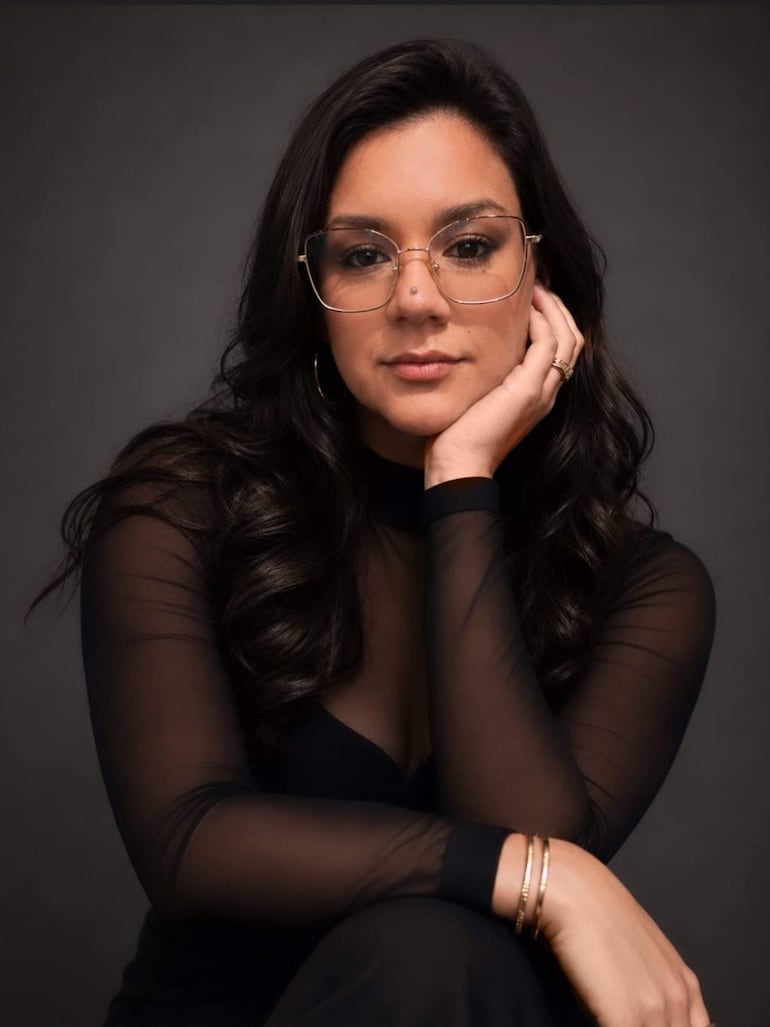 Mujer con cabello largo y ondulado, gafas delgadas y blusa negra, sentada con expresión seria en un entorno oscuro.
