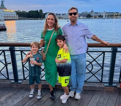 Antonella Volpe en vestido verde sonríe junto a dos niños y un hombre con camisa de cuadros en un muelle al atardecer.