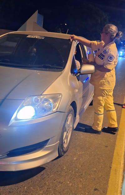Mujer con uniforme amarillo hablando con un conductor en un vehículo plateado, ambiente urbano con luces de vehículos al fondo.
