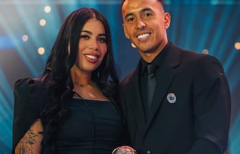 Kaku y Carrizo sonríen en evento formal; ambos visten de negro, Kaku sostiene un trofeo brillante.