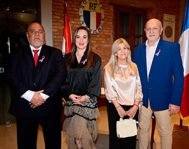 Grupo de cuatro personas en evento formal, sonriendo y con cintas de colores en sus prendas, decorado de ladrillos y bandera en el fondo.