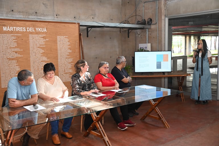 Mujer de pie con micrófono en overol de mezclilla, junto a un panel de madera con nombres y un público atento en ambiente formal.
