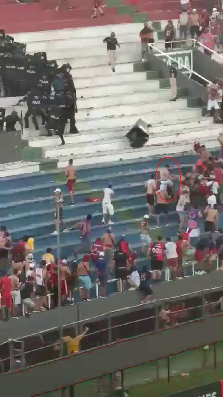 Ambiente de estadio con espectadores agitados. Agentes de policía visibles controlando la situación entre la multitud.