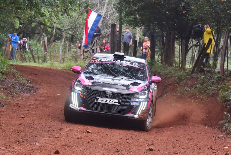 Coche de rally de colores brillantes avanzando velozmente mientras espectadores sostienen una bandera de Paraguay.