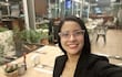 Mujer joven con gafas rosa y blazer negro sonríe en un moderno café, rodeada de decoraciones y plantas.