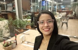 Mujer joven con gafas rosa y blazer negro sonríe en un moderno café, rodeada de decoraciones y plantas.