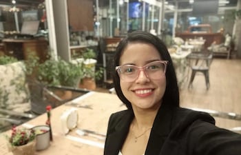Mujer joven con gafas rosa y blazer negro sonríe en un moderno café, rodeada de decoraciones y plantas.