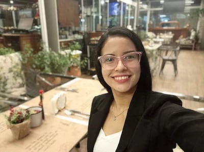 Mujer joven con gafas rosa y blazer negro sonríe en un moderno café, rodeada de decoraciones y plantas.