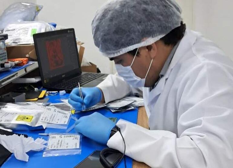 Técnico en laboratorio con bata blanca y guantes azules, concentrado en dispositivos en la mesa azul junto a una laptop.