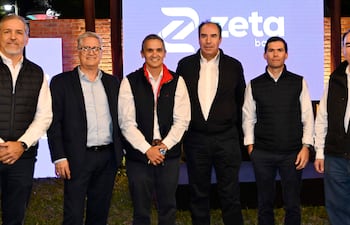 Seis hombres posan en un evento de Zeta Banco, todos con camisas blancas y chalecos oscuros, bajo una pantalla iluminada con el logo 'zeta'.