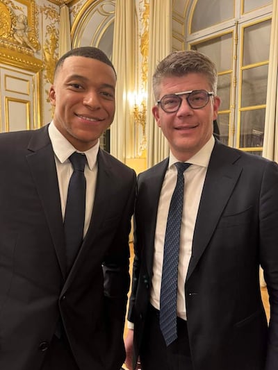 Cedric Perrin y Kylian Mbappé sonríen en trajes oscuros, en un lujoso ambiente decorado con elementos dorados.