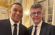 Cedric Perrin y Kylian Mbappé sonríen en trajes oscuros, en un lujoso ambiente decorado con elementos dorados.