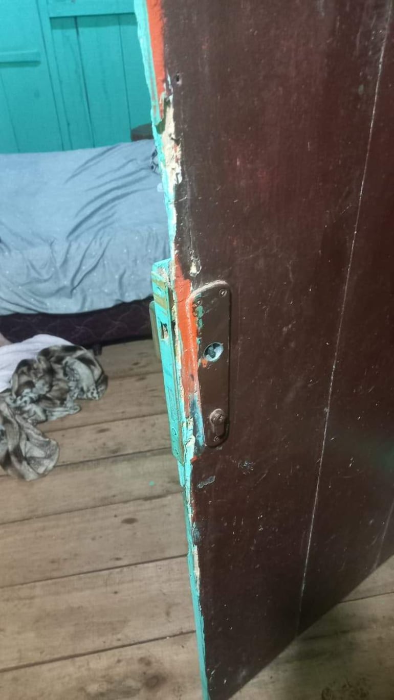 Puerta deteriorada en un ambiente interior, con detalles de pintura desgastada y una cama visible detrás.