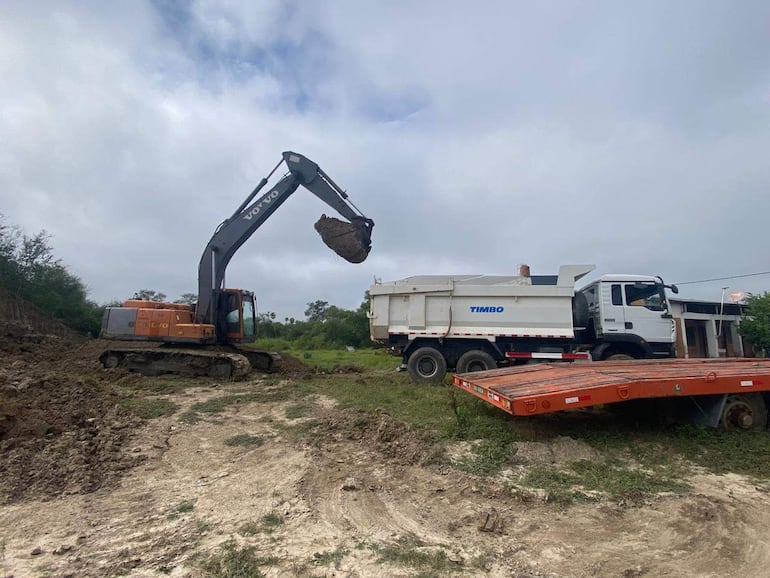 Excavador Volvo y camión de volcado blanco en obra de construcción, rodeados de vegetación y cielo nublado.