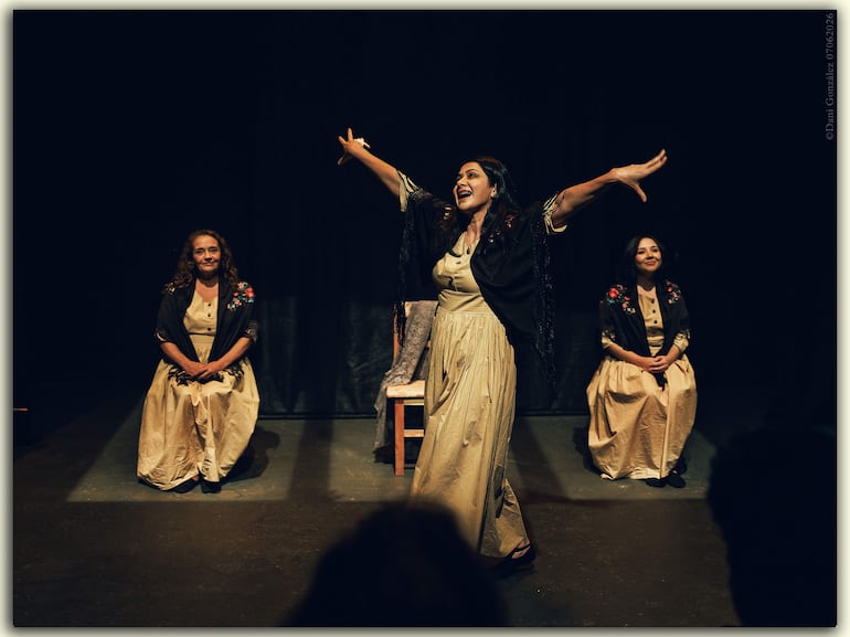 Tres mujeres en un escenario oscuro; la central de pie con atuendo claro y châle negro, levantando los brazos.