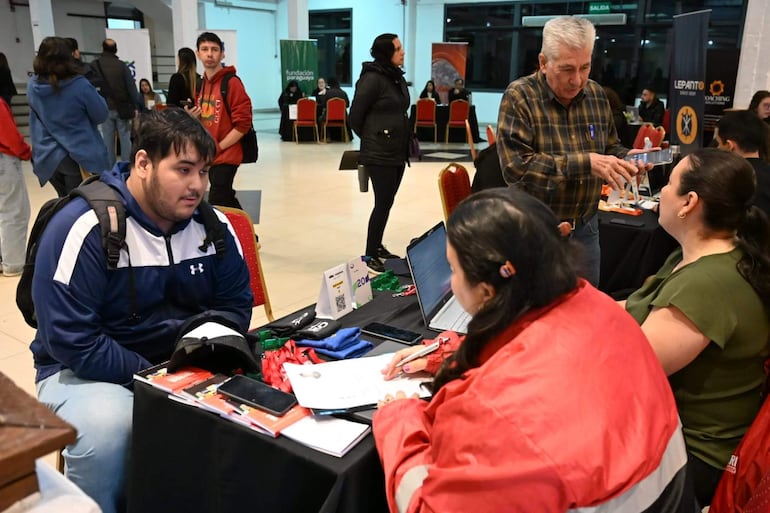 Joven con mochila negra y camiseta azul, observa a mujer con camiseta roja tomando notas, rodeados de un ambiente activo.