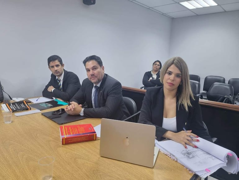 Óscar Tuma con traje oscuro y corbata, Maricel Orihuela en blazer negro, en sala con mesa y documentos.