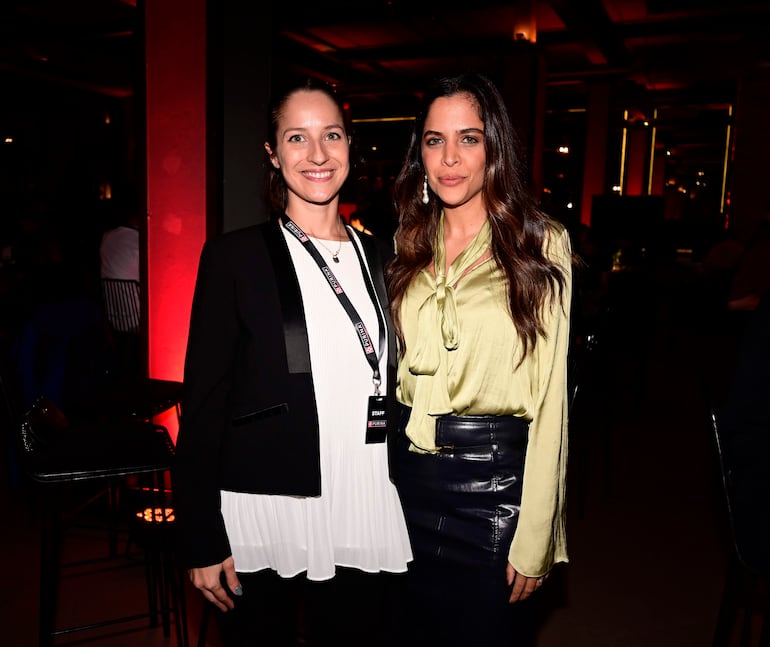 Dos mujeres, una con blusa blanca y chaqueta negra y otra con blusa verde y falda negra, sonríen en un ambiente elegante con luces rojas.