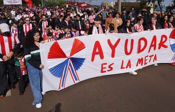 Multitud en camisetas a rayas rojas y blancas, sosteniendo pancarta 'AYUDAR ES LA META', rostros sonrientes y ambiente festivo.