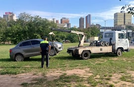 Agente de tránsito con uniforme verde y amarillo vigila un remolque de un coche gris en un parque urbano.