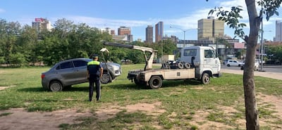 Agente de tránsito con uniforme verde y amarillo vigila un remolque de un coche gris en un parque urbano.