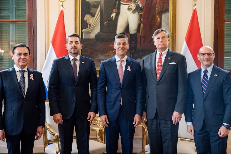 Santiago Peña en traje claro con corbata rosa hablando mientras otros políticos en traje oscuro lo observan atentamente. Banderas de Paraguay visibles.