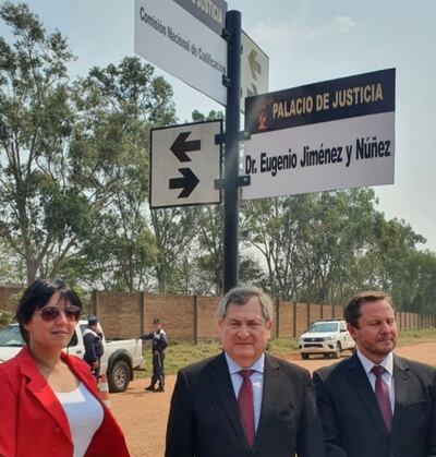 Tres personas frente al Palacio de Justicia; mujer con abrigo rojo, hombre en chaqueta negra con corbata y otro con traje oscuro, ambiente formal.