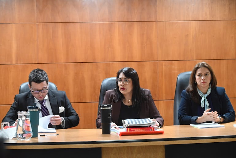 Hombre con traje negro y gafas revisa documentos, flanqueado por dos mujeres en un entorno formal de tribunal.