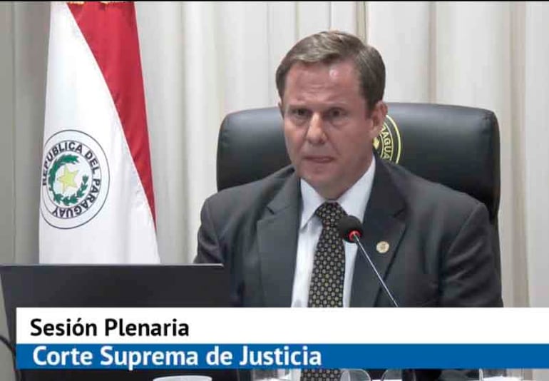 Alberto Martínez Simón en traje oscuro y corbata, sentado en reunión con fondo de bandera paraguaya.