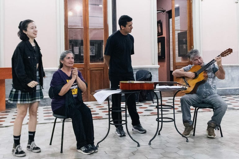 Hombre canoso tocando guitarra, mujer de camiseta morada escuchando atenta, joven de camiseta negra observando, y mujer con abrigo negro al fondo.