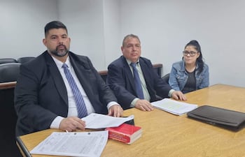 Nery Villalba con barba y traje oscuro, Ricardo Paredes en camisa clara y saco oscuro, Ada Arasy Ruiz Díaz con gafas en chaqueta de mezclilla, sentados en mesa con documentos.