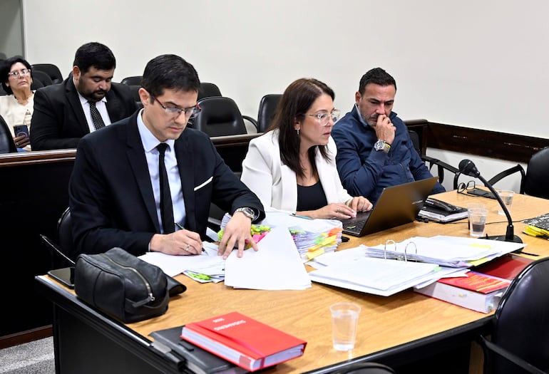 Grupo de tres personas en mesa: hombre con gafas escribiendo, mujer con computadora y hombre pensativo, en sala formal.