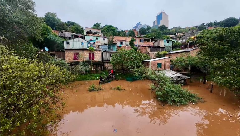 Cuatro personas en un bote entre aguas marrones, rodeadas de vegetación y edificios, reflejando una situación de emergencia.