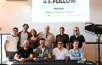 Once personas en un evento, algunos sentados sosteniendo un megáfono y un clapboard, luciendo felices en un ambiente decorado con logos.