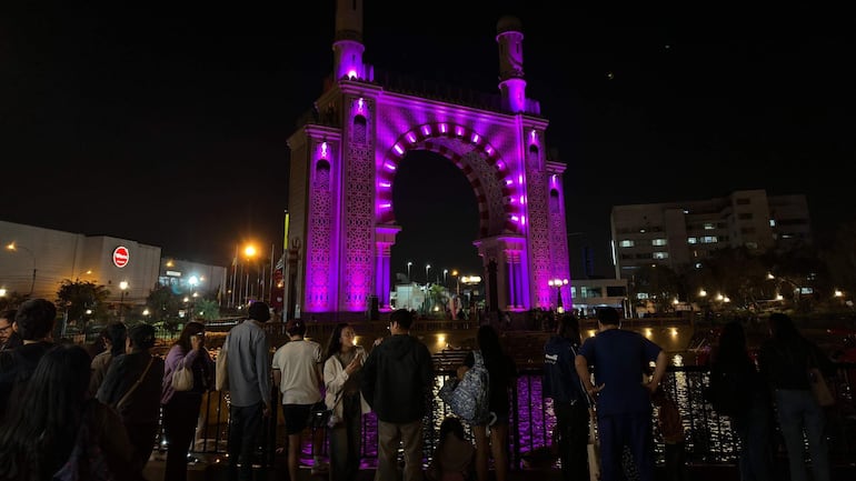 Multitud alegre frente a arco decorativo iluminado en morado; personas conversan y disfrutan del ambiente festivo nocturno.