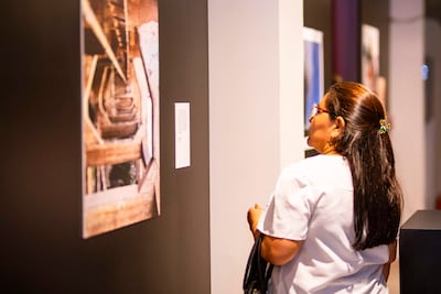 Mujer de perfil con cabello largo y lacio, blusa clara y gafas, observando una fotografía enmarcada en la galería.