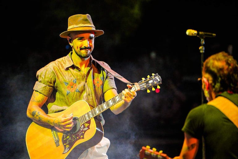 Hombre sonriente tocando guitarra acústica con camisa clara y sombrero, mientras otro con camiseta negra lo acompaña de espaldas.