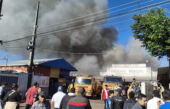 Multitud preocupada observa humo negro del incendio en el Mercado Municipal de Itauguá, con camiones de bomberos amarillos de fondo.