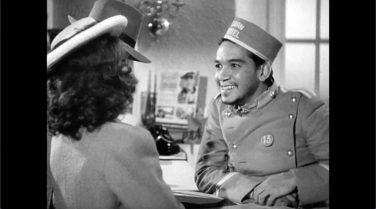 Mario Moreno sonriente, vestido como Cantinflas, conversa con una mujer con cabello rizado y sombrero en un hotel.