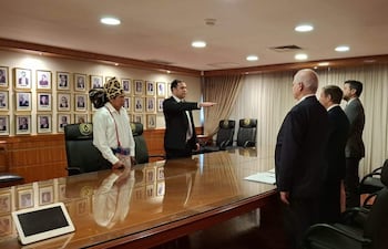Hombre en traje jurando con la mano levantada, rodeado de hombres en trajes oscuros y un representante indígena con tocado.