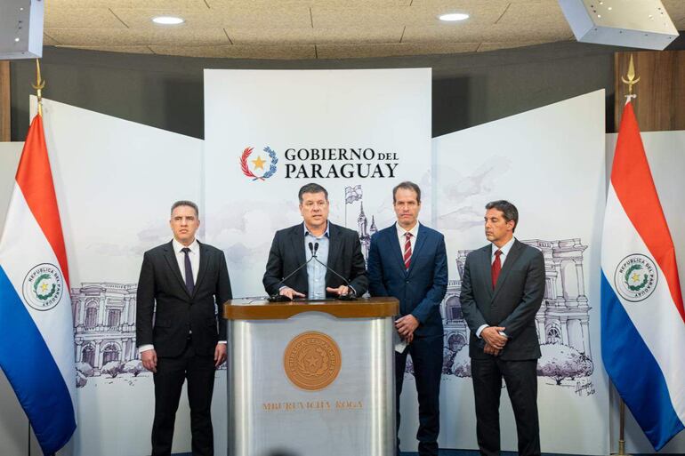 Gustavo Villate en traje oscuro escuchando a Javier Giménez, quien habla con micrófono. Fondo con banderas del Paraguay.