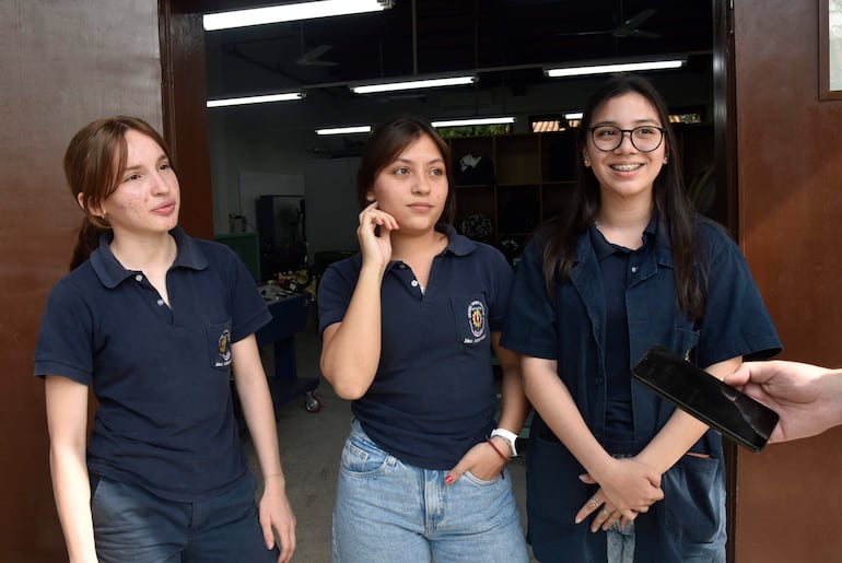 Tres jóvenes en camisetas azules discuten en taller, una sonriente con gafas, otra pensativa, en un ambiente iluminado.