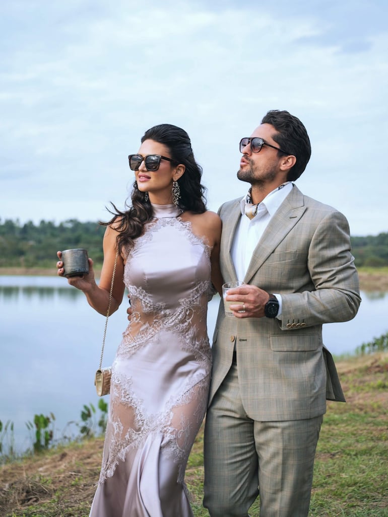 Leryn Franco en vestido claro y Rodrigo Galeano en traje claro, ambos usando gafas de sol y sosteniendo vasos, rodeados de vegetación.