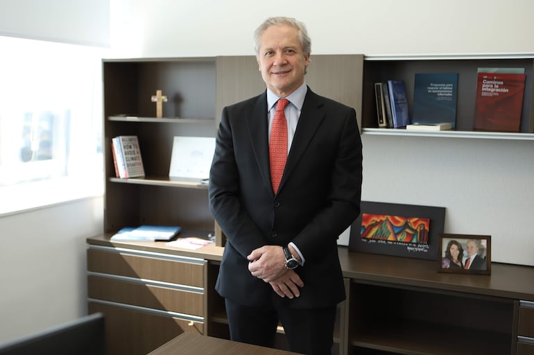 Jorge Srur, con traje negro y corbata roja, de pie en una oficina con estantes de libros y objetos decorativos detrás.